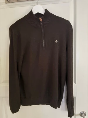 Morris zip - Snygg mörkbrun stickad tröja med halv dragkedja och ribbad hög krage. Tröjan har en liten broderad detalj på bröstet och ribbade muddar vid ärmslut och nederkant. Perfekt för lager på lager och en stilren look. Skick 9/10