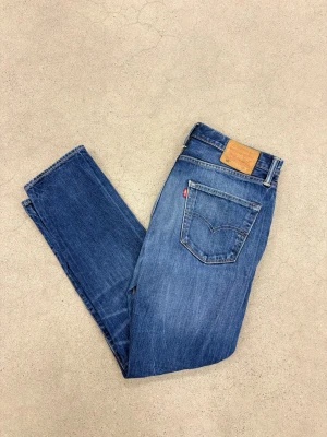 Blå jeans Levi's 32/32 - Klassiska blå jeans från Levi's med rak passform och fem fickor. Jeansen har en snygg tvättad look, läderpatch bak i midjan och röda Levi's-detaljer vid bakfickan. Tillverkade i slitstark denim, perfekta för en avslappnad stil.                        Storlek W32 L33✅