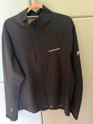 Svart regnjacka Peak Performance - Snygg svart regnjacka från Peak Performance med dragkedja framtill och diskreta loggor på bröstet och ärmen. Jackan har minimalistisk design, hög krage och justerbar nederkant. Perfekt för regniga dagar och outdoor-äventyr.