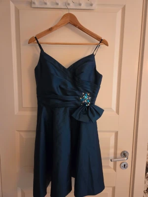 Marinblå kort klänning med rosett - Snygg marinblå klänning med smala axelband och v-ringning. Klänningen har en dekorativ rosett och glittrande pärldetaljer i midjan. Tillverkad i ett glansigt material som ger en elegant look. Perfekt för speciella tillfällen.