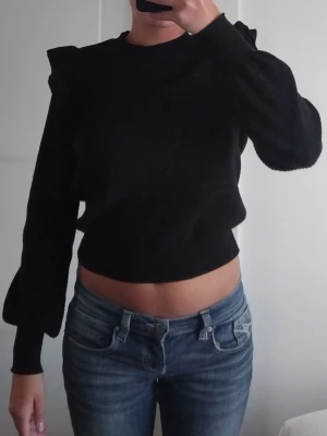 Svart stickad tröja med volangdetaljer - Trendig svart stickad tröja med volangdetaljer vid axlarna och ballongärmar. Tröjan är ribbad och en något croppad passform. Står M på lappen men passar perfekt på S också.