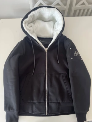 Svart Moose Knuckles Bunny med vit fleecefoder - Snygg svart Bunny från Moose Knuckles med dragkedja, två stora fickor framtill och vit fluffig fleece på insidan av huvan. Ärmen har en svart logodetalj i metall på en patch. Perfekt för dig som vill ha en varm och stilren hoodie.
