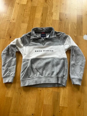 Gråvit zip tröja Race Marine - 146/152cl. Säljer en stilren grå och vit sweatshirt från Race Marine med halv dragkedja och tryckt logga på bröstet. Tröjan har ribbade muddar och sportig design, perfekt för en avslappnad look. Materialet är mjukt och bekvämt. Nyskick, bara provad.