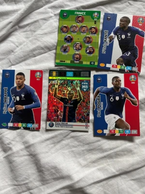 Fotbollssamlarspelkort Frankrike - Säljer fem fotbollssamlarspelkort med franska landslagsspelare. Korten har färgstarka motiv med spelare som Kylian Mbappé, N'Golo Kanté, Paul Pogba och Blaise Matuidi. Korten är i klassisk rektangulär form och gjorda av glansigt papper.