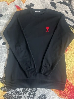 Ami Paris  - Snygg svart stickad tröja med rund hals och ett broderat rött hjärta med bokstaven A på bröstet. Tröjan har ribbade muddar vid ärmslut och nederkant. Perfekt för dig som gillar stilrena plagg med en twist.