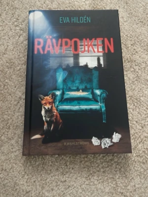 Rävpojken av Eva Hildén - Boken 'Rävpojken' av Eva Hildén har ett omslag med en röd räv och en turkos fåtölj mot en mörk bakgrund. Omslaget har även detaljer som hopknycklat papper och fönster med persienner. Utgiven av B. Wahlströms. Det finns även ett litet märke på framsidan som man ser tydligare på den 2a bilden.☺️🩷