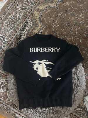 Svart stickad Burberry-tröja - Snygg svart stickad tröja från Burberry med vit logga och ikoniskt riddarmotiv på bröstet. Tröjan har rund halsringning, långa ärmar och ribbade muddar. Perfekt för dig som vill ha en statement-piece i garderoben.