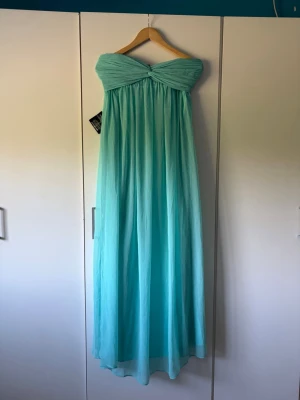 Turkos balklänning  - Säljer en ljusblå axelbandslös maxiklänning med draperad överdel och flytande kjol i tunt, skirt tyg. Perfekt för dig som vill ha en elegant och luftig look. Klänningen har en romantisk känsla och passar till speciella tillfällen.