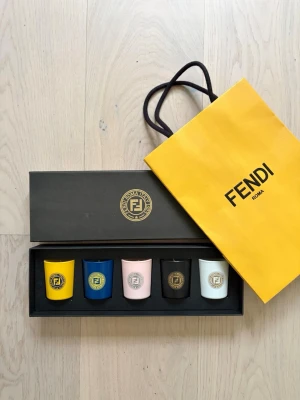 Fendi doftljus set i presentförpackning - Unused set of five scented candles in the original Fendi presentation box. Featuring the elegant signature fragrance used in Fendi boutiques.  Includes: • 5 unused candles • Original Fendi box • Fendi shopping bag (included as a gift)  Condition: New and unused. The outer box has a few minor marks from storage on the front, as shown in the photos.