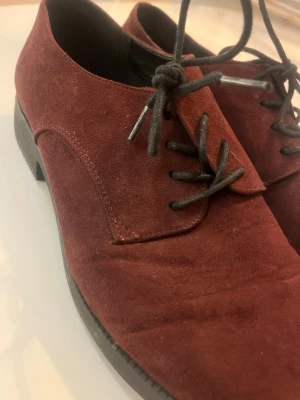 Vinröda mockaloafers med snörning - Säljer ett par vinröda loafers i mocka med svarta snören och låg klack. Skorna har en klassisk, stilren design och passar perfekt till dig som vill ha något snyggt och bekvämt. Materialet är mjukt och ger en lyxig känsla.