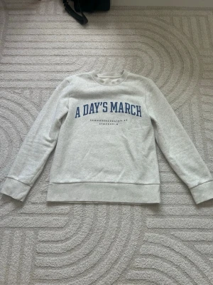 Grå sweatshirt A Day's March - Snygg ljusgrå sweatshirt från A Day's March med blå text på bröstet och rund hals. Tröjan har ribbade muddar vid ärmslut och nederkant. Perfekt för en avslappnad och stilren look. Hör av sig vid frågor.