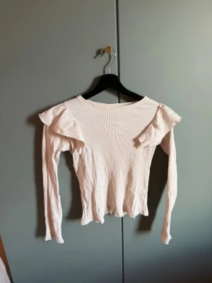 Ljusbeige ribbad topp med volang - Säljer en ljusbeige ribbad topp med långa ärmar och volangdetaljer vid axlarna. Toppen har en figurnära passform och är tillverkad i ett mjukt, stretchigt material som känns bekvämt mot huden. Perfekt för dig som gillar feminina detaljer och enkel stil.