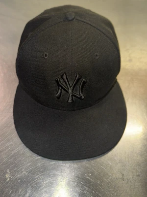 Svart New York Yankees keps - Säljer en svart keps med New York Yankees-logga framtill, broderad ton-i-ton. Klassisk flat peak och ventilerande hål upptill. Perfekt för dig som gillar streetwear och sportig stil.