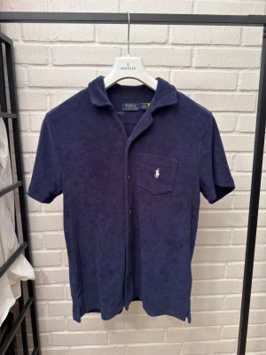 Marinblå frottéskjorta från Polo Ralph Lauren - Säljer en marinblå skjorta i mjuk frotté från Polo Ralph Lauren. Skjortan har korta ärmar, klassisk krage, knappar framtill och en bröstficka med den ikoniska broderade loggan. Perfekt för en chill och avslappnad stil.