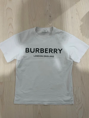 Vit Burberry t-shirt med tryck - Vit t-shirt från Burberry med klassiskt svart logotyp-tryck på bröstet och texten 'London England' under. T-shirten har rund halsringning och korta ärmar. Tillverkad i mjuk bomull för en skön känsla och clean look.