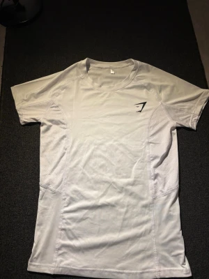 Gymshark compression shirt   - Säljer en vit kortärmad träningströja från Gymshark. Tröjan har rund halsringning, ventilerande meshpaneler på sidorna och en diskret svart Gymshark-logga på bröstet. Perfekt för gymmet eller löprundan. Aldrig använd, sitter mer som S.
