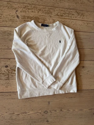 Vit sweatshirt från Polo Ralph Lauren - Klassisk vit sweatshirt från Polo Ralph Lauren med broderad svart logga på bröstet. Tröjan har rund halsringning, långa ärmar och ribbade muddar. Perfekt för en clean och stilren look.