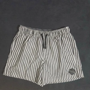 PP badbyxor - Snygga badbyxor med svartvita vertikala ränder och elastisk midja med dragsko. På vänster ben finns en svart patch med PP-logga i silver. Perfekta för stranden eller poolen, med klassisk och stilren look.