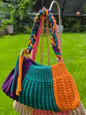 Färgglad virkad handväska med flätat handtag - Unik handväska i virkade sektioner med lila, turkos, orange och rosa färger. Väskan har ett flätat handtag i matchande färger och en guldfärgad kedja som axelrem. Materialet är tjockt garn som ger en mjuk och trendig look. Perfekt för dig som vill sticka ut med accessoarer.