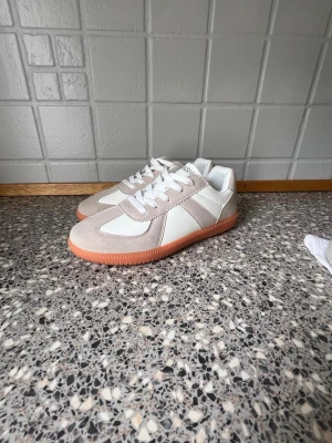 Vita och beige sneakers med gummisula - Snygga sneakers i vitt och beige med klassisk låg siluett. Ovandelen är i en mix av mocka och slätt material, och skorna har vita snören samt en kontrasterande ljusbrun gummisula. Perfekta för en clean och stilren look.
