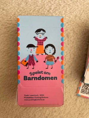 Spelet om Barndomen sällskapet spel - Kortspel med titeln 'Spelet om Barndomen' från Pushing Buttons. Färgglad ask i blått, rosa och orange med tecknade figurer på framsidan. Innehåller spelkort med illustrationer och namn. Perfekt för spelkvällar och samtal.