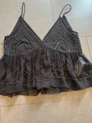 Svart pärlbroderad linnetopp Gina Tricot - Supersnygg svart linnetopp från Gina Tricot med tunna axelband och v-ringning. Toppen har pärlbroderade detaljer i geometriska mönster och är tillverkad i ett tunt, lätt material som ger en luftig känsla.