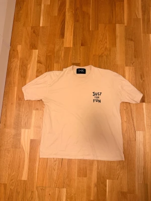 Beige t-shirt med popcorntryck medium - Beige t-shirt med trycket 'JUST FOR FUN' fram och ett stort popcornmotiv med texten 'GET SOME POPCORN' på ryggen. Skön passform och klassisk rund hals. Perfekt för dig som gillar roliga prints och chill stil. Medium