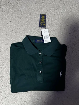 Mörkgrön pikétröja från Ralph Lauren - Klassisk mörkgrön pikétröja från Ralph Lauren med vit broderad logga på bröstet. Tröjan har krage, knappar framtill och korta ärmar. Perfekt till både jeans och shorts för en clean och stilren look.