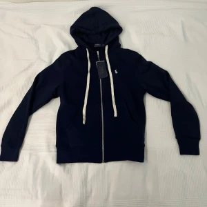 Marinblå hoodie från Polo Ralph Lauren - Snygg marinblå hoodie med dragkedja från Polo Ralph Lauren. Hoodien har vit broderad logga på bröstet, vita dragsnören och en klassisk huva. Perfekt för en avslappnad och stilren look. Mjuk bomullskvalitet och ribbade muddar.