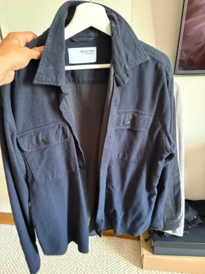 Mörkblå overshirt från Selected Homme - Stilren mörkblå overshirt från Selected Homme med två stora bröstfickor med knappstängning och klassisk krage. Skjortan har långa ärmar och knappar framtill. Perfekt lager-på-lager-plagg i mjukt material.