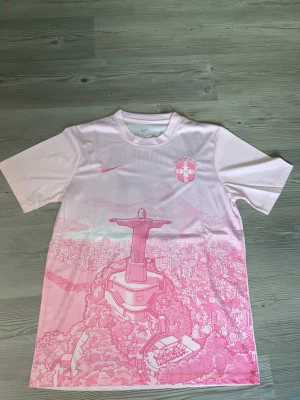 Rosa Neymar Jr fotbollströja - Säljer en rosa fotbollströja med Neymar Jr och nummer 10 på ryggen. Framsidan har ett tryck av Kristusstatyn i Rio och stadsmotiv i rosa toner. Tröjan har korta ärmar och rund hals, samt Nike-logga och klubbmärke på bröstet.