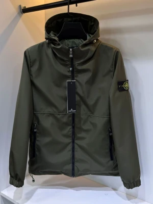 Stone Island Windbreaker  - Snygg olivgrön vindjacka från Stone Island med huva och dragkedja framtill. Jackan har två sidofickor med dragkedja och den klassiska logotypen på vänster ärm. Perfekt för en streetwear-look och enkel att matcha med olika outfits.