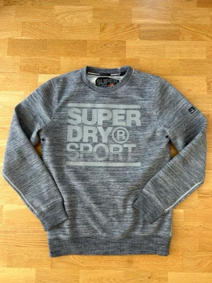Grå sweatshirt från Superdry Sport - Snygg gråmelerad sweatshirt från Superdry Sport med stort tryck på bröstet. Tröjan har rund halsringning, ribbade muddar och diskreta detaljer på ärmarna. Perfekt för en sportig och avslappnad stil.