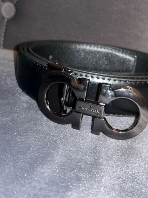 Salvatore Ferragamo - Snyggt svart skärp från Salvatore Ferragamo med ikoniskt dubbel-Gancini-spänne i silverfärgad metall. Bältet är gjort i slätt skinn och har en stilren, klassisk look som passar till många outfits. Perfekt accessoar för dig som gillar exklusiva detaljer.