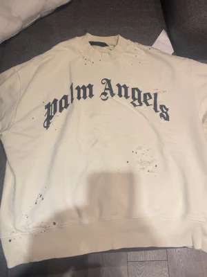 Vit hoodie från Palm Angels - Säljer en oversized vit hoodie från Palm Angels med stor svart logga i gotisk stil över bröstet. Hoodien har unika slitningar och färgstänk som ger en edgy streetwear-look. Tillverkad i mjukt bomullsmaterial och har ribbade muddar vid ärmslut och nederkant.