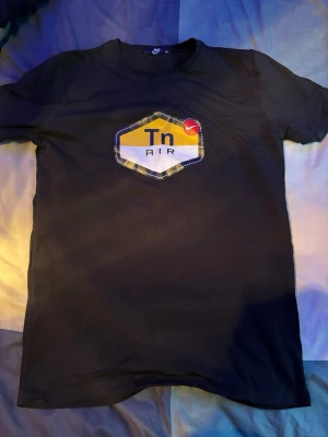Svart Nike Tn Air t-shirt - Svart t-shirt från Nike med Tn Air-logga i gult, vitt och blått på bröstet. Klassisk rund hals och korta ärmar. Perfekt för dig som gillar streetwear och sportig stil. T-shirten passar även som S eftersom den är en tajt M storlek passform.