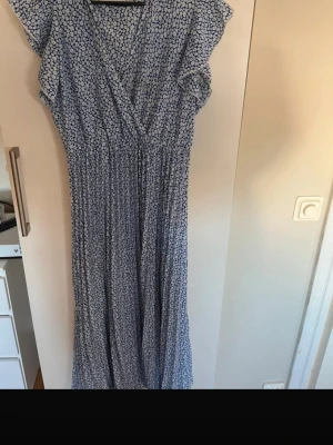 Blåvit blommig omlottklänning - Söt blåvit omlottklänning med småblommigt mönster och korta volangärmar. Klänningen har en markerad midja och faller i en luftig, vadlång modell. Perfekt för vår och sommar, tillverkad i ett lätt och skönt tyg.