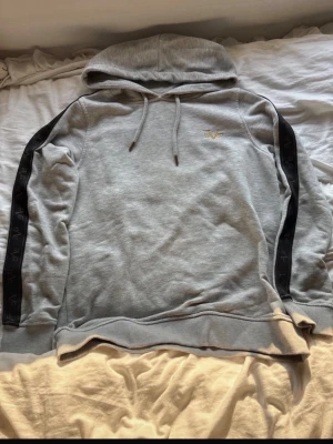 Grå hoodie med svarta detaljer - Snygg grå hoodie från V1969 med broderad logga i guld på bröstet. Hoodien har svarta band med logga längs ärmarna och justerbar huva med dragsko. Perfekt för en avslappnad och trendig stil.