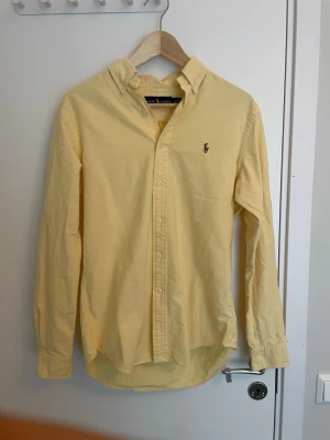 Gul skjorta från Ralph Lauren - Klassisk gul skjorta från Ralph Lauren med button-down krage och broderad logga på bröstet. Skjortan har långa ärmar och knäppning framtill. Perfekt för en stilren och avslappnad look. Pris kan självklart diskuteras. 