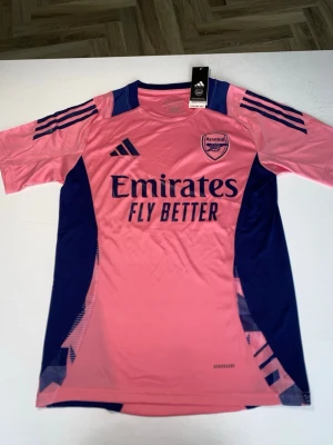 Arsenal rosa matchtröja från Adidas - Säljer en rosa Arsenal matchtröja från Adidas med mörkblå detaljer och logga på bröstet. Tröjan har korta ärmar, rund hals och är tillverkad i ett lätt och ventilerande polyestermaterial. Perfekt för fotbollsträning eller att bära som supporter.