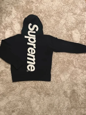 MÖRKBLÅ Supreme hoodie med stor logga på baksidan - Supreme hoodie i MÖRKBLÅ med stor vit Supreme-logga broderad vertikalt längs ryggen och huvan. Klassisk streetwear-look med huva och ribbade muddar. Perfekt för dig som vill sticka ut med en ikonisk design.