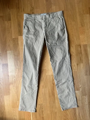 Produkt beige chinos med raka ben - Snygga beige chinos med raka ben och klassisk passform. Byxorna har sidofickor fram och två bakfickor med knapp. Tillverkade i ett mjukt bomullsmaterial som känns skönt mot huden. Perfekta för en clean och avslappnad stil.