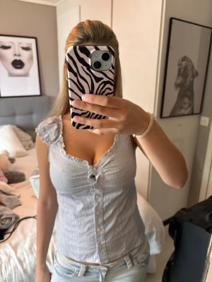 Randig blus med volangdetalj - Säljer en söt blus med smala vertikala ränder i vitt och ljusblått. Toppen har volangdetaljer vid axlarna, v-ringning och knappar framtill. Ärmlös och figurnära passform, perfekt för varma dagar. Storlek Xs-S