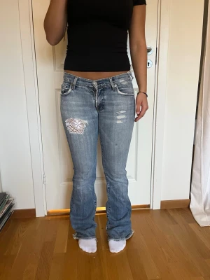 Ljusblå bootcut jeans med spetsdetalj - Säljer ett par lågmidjade ljusblå bootcut jeans från 7 for all Mankind med spetsdetalj på vänster lår. Gamla slitningar/hål är helt lagade från insidan och syns inte utifrån. Pris går att diskutera