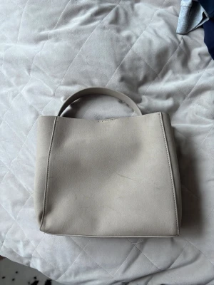 Beige handväska Gina tricot - Snygg beige handväska i minimalistisk design med bredt handtag och rak form. Väskan har ett innerfack med dragkedja för småsaker och är gjord i slätt konstläder. Perfekt för dig som gillar enkel och modern stil.