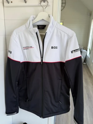 Porsche Motorsport vindjacka från BOSS - Snygg vindjacka i svart och vitt med röda detaljer från BOSS och Porsche Motorsport. Jackan har dragkedja framtill, hög krage och loggor från Mobil 1 och Michelin på ärmarna. Perfekt för dig som gillar racing och sportig stil.