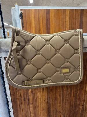 Beige schabrak från Equestrian Stockholm - Säljer ett snyggt beige schabrak från Equestrian Stockholm med quiltat mönster och guldfärgade detaljer. Schabraket har en elegant kant i tvinnat snöre och en metallbricka med logga. Perfekt till hästutrustningen! Nypris:900kr 