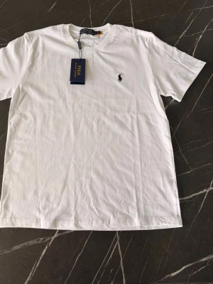 Vit t-shirt från Polo Ralph Lauren - Klassisk vit t-shirt från Polo Ralph Lauren med den ikoniska lilla loggan broderad på bröstet. Tillverkad i mjuk bomull och har en normal passform med korta ärmar och rund hals. Perfekt basplagg till garderoben.