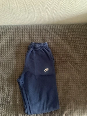 Blåa mjukisbyxor från Nike - Säljer ett par blåa mjukisbyxor från Nike med vit logga på vänster ben. Byxorna har resår i midjan och muddar vid bensluten. Perfekta för chill dagar eller träning. Tillverkade i mjukt och bekvämt material.