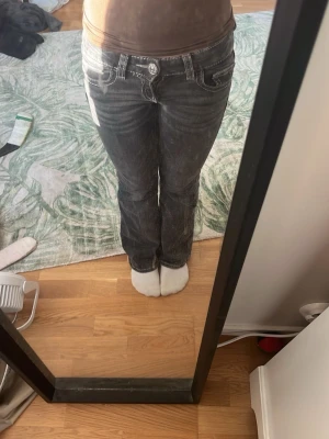 Svarta bootcut jeans med kontrastsömmar - Säljer ett par svarta bootcut jeans med vita kontrastsömmar och dekorativa fickor bak. Jeansen har låg midja och klassisk femficksdesign. Snygga detaljer med knappar på bakfickorna och slitningar för en cool look.